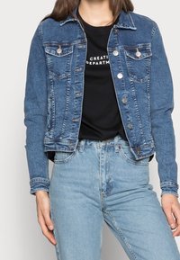 Denimjacka i mörkblått, med knappstängning fram, två bröstfickor och en något kortad design. Matchad med en svart t-shirt och ljusa jeans.