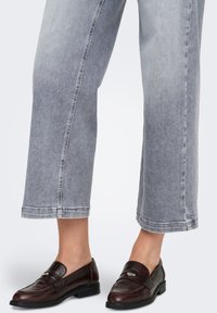 Rjavi usnjeni loaferji z zaobljenim sprednjim delom in srebrnimi okrasnimi elementi, kombinirani z zložkami na širokih sivih kavbojkah. Gladek tekstura in poliran zaključek.