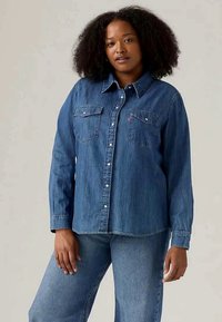 Camisa de mezclilla en azul oscuro con manga larga, cierre de botones en la parte delantera, dos bolsillos en el pecho y un cuello puntiagudo. Textura suave y corte relajado.
