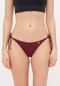 Burgundy bikini bottom with ribbed texture, adjustable side ties, and a small metallic accent tag. Design is low-rise and minimalistic.