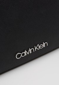 Lähikuva mustasta, teksturoidusta pinnasta, jossa on hopeanvärinen metallinen "Calvin Klein"-logo kiinnitettynä alaoikealle.