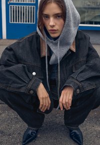 Giacca in denim nera oversize con felpa grigia con cappuccio sotto. Il modello è accovacciato sul marciapiede, mostrando scarpe nere appuntite e maniche dettagliate.