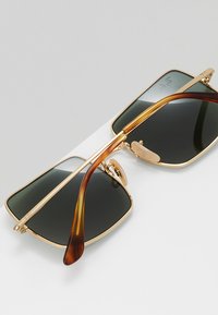 Occhiali da sole con montatura in metallo dorato e lenti verde scuro, caratterizzati da aste degradé marroni e un design rettangolare elegante.