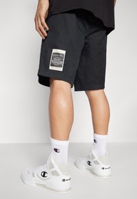 Pantalones cortos negros con un parche de logo en el costado, combinados con calcetines blancos y zapatillas blancas con logo. El fondo es liso.