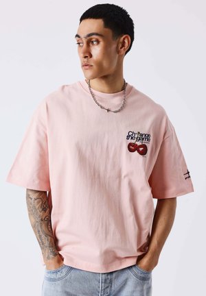 Jeune homme aux cheveux courts et foncés portant un t-shirt rose avec un motif cerise et le texte « Change the game », mains dans les poches, regardant à gauche.