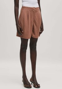 Braune gefaltete Shorts aus weichem Stoff, mit seitlichen Taschen und einem lässigen Schnitt. Getragen mit schwarzen Sandalen mit Riemen.