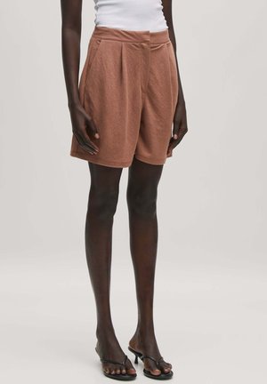 Bruine geplooide shorts gemaakt van zacht stof, met zijzakken en een relaxte fit. Draagt met zwarte sandalen met banden.