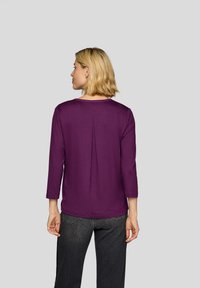 Lila Langarmshirt mit rundem Ausschnitt und Plissee-Detail am Rücken, kombiniert mit dunkler Jeans. Glatte Stoffstruktur.