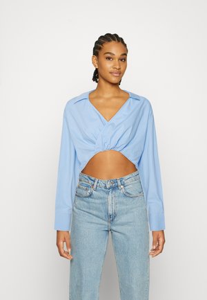 Camisa cropped azul claro com decote em V e frente franzida, combinada com jeans de denim claro de cintura alta. Mangas longas com punhos de botão.