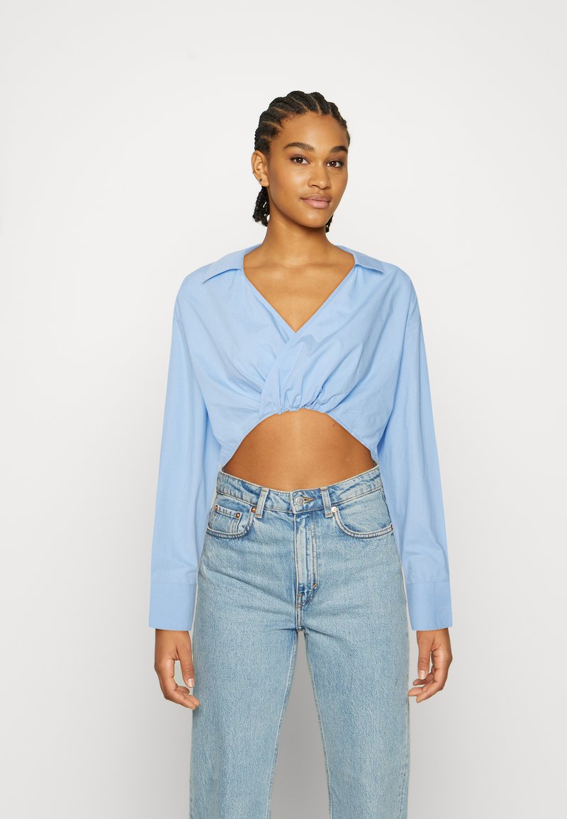 Camisa cropped azul claro com decote em V e frente franzida, combinada com jeans de denim claro de cintura alta. Mangas longas com punhos de botão.