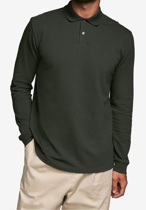 Homme portant un polo à manches longues vert foncé avec deux boutons et un pantalon beige clair, les mains détendues le long du corps.