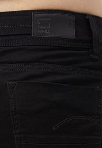 Vaqueros de denim negro con un parche de cuero con el logo grabado, costuras visibles y un único bolsillo trasero con un sutil detalle de costura curva.