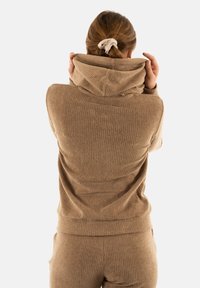 Sudadera de punto beige con cuello alto. Textura suave, diseño ajustado y un bolsillo tipo canguro. Llevada con pantalones a juego de punto.