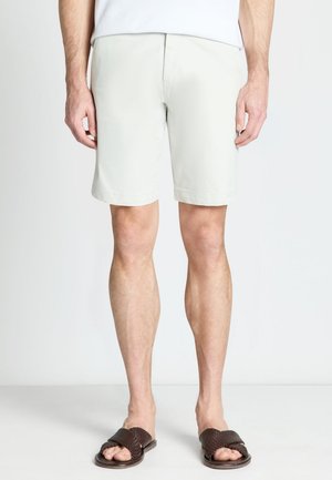Homme portant un short beige clair et des sandales tressées marron foncé, debout devant un fond clair uni.