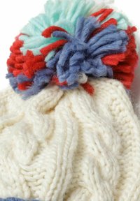 Weißer Strickbeanie mit Kabelmuster, gekrönt von einem mehrfarbigen Pompon aus roten, blauen und mintgrünen Garnen.