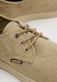 Zapatos de lona beige con cordones redondos, ojales de metal y una etiqueta que dice “INSIDE”, con superficies lisas y texturizadas.