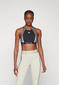 Svart og grå sports-BH med racerback-design, med et elastisk bånd merket "TECHFIT." Sammen med høyhøpet lysegrå tights.