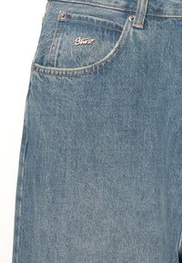 Primo piano di jeans in denim blu che mostra la tasca anteriore con rivetti metallici e una piccola spilla argentata del marchio "Swiz".