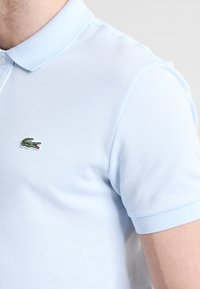 Svetlo modra polo majica, izdelana iz teksturirane tkanine, s ovratnikom in vezenim zelenim krokodilom na levi prsni strani.