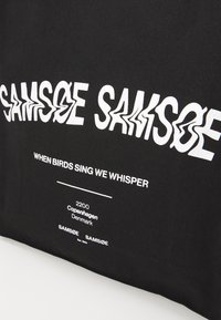 Black canvas bag featuring distorted white text that reads "SAMSOE SAMSOE" and "WHEN BIRDS SING WE WHISPER," with location details below.
