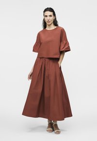 Femme portant une tenue deux pièces couleur rouille avec un haut carré aux manches volantées et une longue jupe plissée, assortie à des sandales blanches à brides.