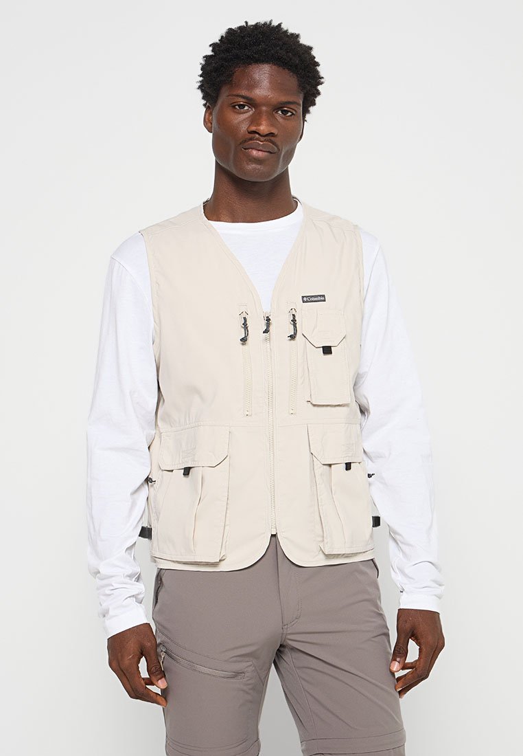 Man in een beige Columbia vest met rits over een wit overhemd met lange mouwen en grijze broek, staande tegen een effen lichte achtergrond.