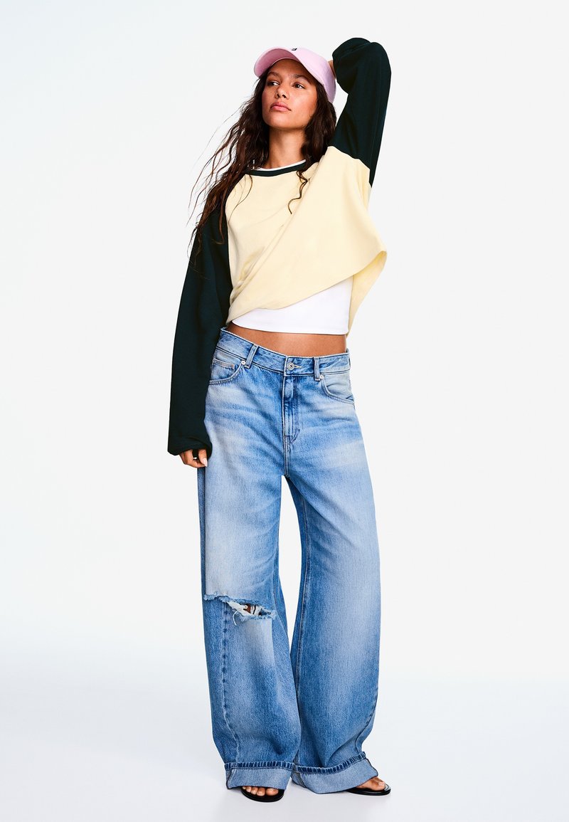 Woman wearing a pink cap, oversized two-tone sweatshirt, white crop top, wide ripped jeans, and black sandals standing with one arm raised.
