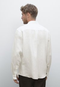 Homme avec les cheveux courts tourné vers la gauche, portant une chemise blanche à manches longues et un pantalon sombre sur un fond gris clair uni.