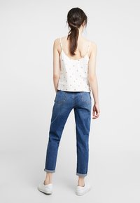 Mujer con el cabello oscuro recogido, vistiendo una blusa blanca de tirantes con estampado floral, jeans azules remangados y zapatillas blancas, de pie, de espaldas.
