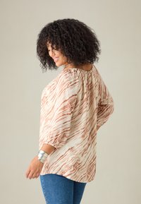 Blusa leggera e stampata con un design ondulato beige e ruggine, scollatura elasticizzata e maniche a sbuffo a tre quarti. Abbinata a jeans blu.
