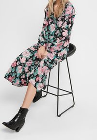 Musta kukallinen mekko, jossa on vaaleanpunaisia ja vihreitä kukkia, pitkät hihat ja helppo design, yhdistettynä mustiin nilkkabuutseihin tuolilla.