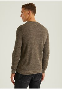 Grau-brauner gestrickter Pullover mit gerippten Bündchen und Saum, mit einem dezenten Logo-Detail am Nacken hinten, kombiniert mit dunklen Jeans.