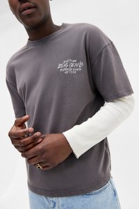 Mand iført lagdelte grå BDG jeans t-shirt over hvid langærmet, lyseblå jeans og en sort ring på venstre hånd.