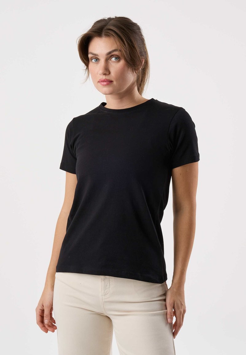 Vrouw met een lichte huid en bruin haar in een staart, gekleed in een strakke zwarte shirt met korte mouwen en lichtbeige broek, staand tegen een effen witte achtergrond.