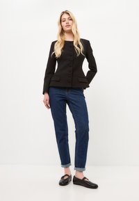 Schwarzer taillierter Blazer mit runden Knöpfen und zwei Fronttaschen, kombiniert mit blauen Jeans mit geradem Bein und hochgekrempelten Ärmeln sowie schwarzen Slippern.