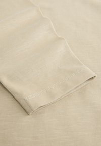 Beežist puuvillast kangas, millel on sile tekstuur, volditud varrukas ja nähtavad õmblused ääres.