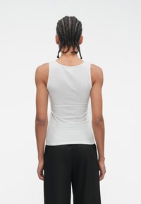 Biały dopasowany top na ramiączkach z okrągłym dekoltem i szerokimi ramiączkami, wykonany z gładkiej bawełny. Model nosi czarne luźne spodnie.