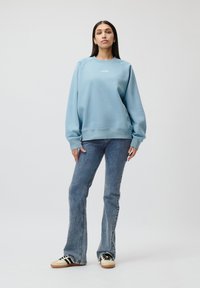 Felpa azzurra chiara con collo rotondo e polsini a costine, abbinata a jeans svasati e sneakers casual con strisce.