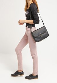 Borsa a tracolla in pelle nera con tasca frontale con zip, forma strutturata e tracolla regolabile. Abbinata a pantaloni fitted rosa chiaro e scarpe nere.