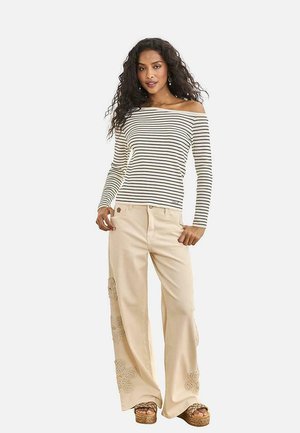 Mujer con el pelo largo y rizado que lleva una blusa de rayas sin hombros, pantalones beige de pierna ancha con bordado floral y sandalias con plataforma.