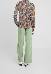 Blusa de manga larga con un estampado floral en negro, blanco y naranja, combinada con pantalones de pana anchos en verde claro y botas de tacón blancas.
