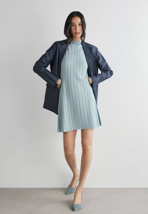 Frau trägt hellblaues Strickkleid, marineblauen Blazer und hellblaue High Heels, steht mit den Händen in den Taschen des Blazers vor einer schlichten Wand.