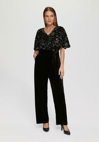 Schwarzes Paillettenoberteil mit V-Ausschnitt und kurzen Ärmeln, kombiniert mit schwarzen samtigen, weit geschnittenen Hosen. Das Modell trägt schwarze High-Heels.