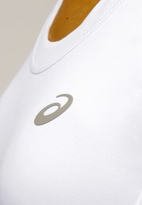 Top atlético blanco sin mangas con un escote redondeado y un acabado texturizado. Presenta un acento de logo plateado en la zona del pecho.