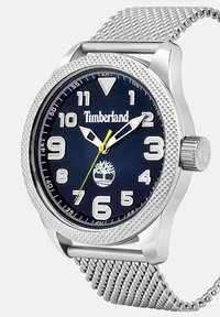 Srebrna ura iz nerjavečega jekla z modrim teksturiranim številčnikom, belimi indeksi in rumeno sekundno kazalcem. Ima mrežasto zadrgo in logotip Timberland.