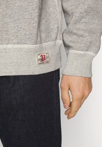 Gray sweatshirt with a textured fabric, ribbed cuffs, and waistband. Features a vintage logo patch on the lower front.