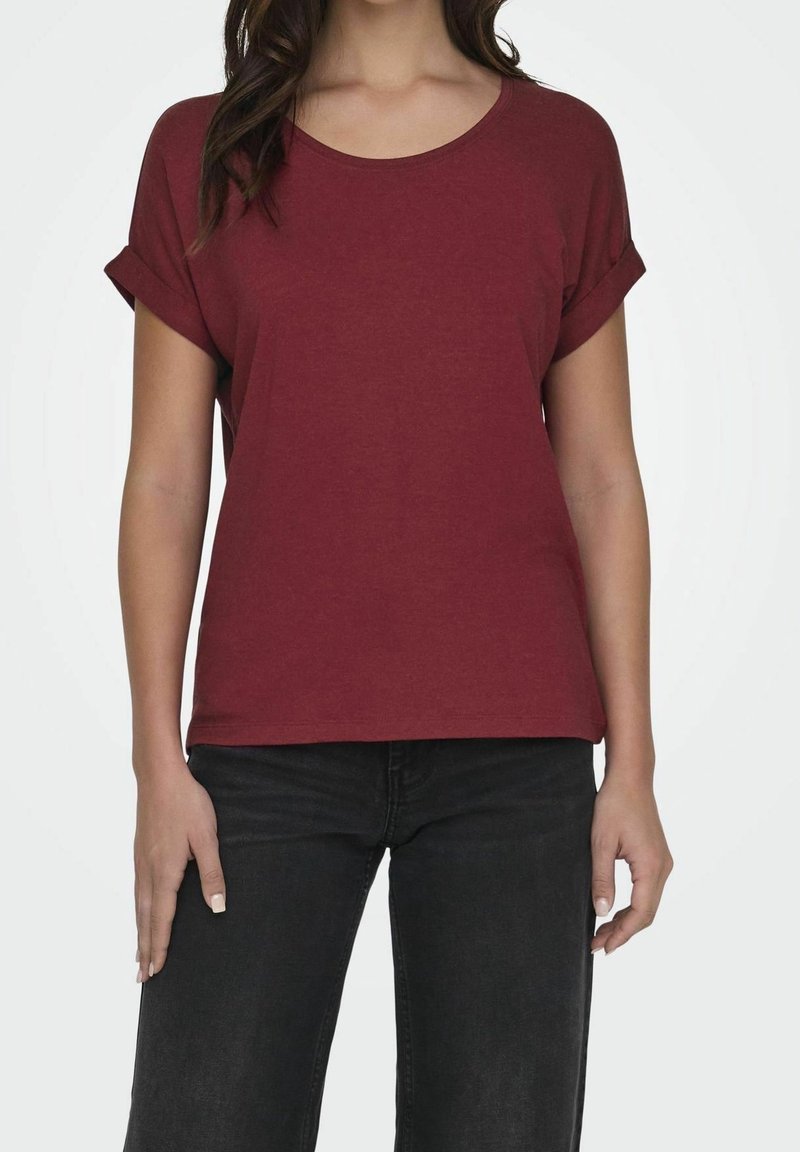 Femme portant un t-shirt bordeaux uni à manches courtes retroussées et un jean noir, debout devant un fond clair uni.
