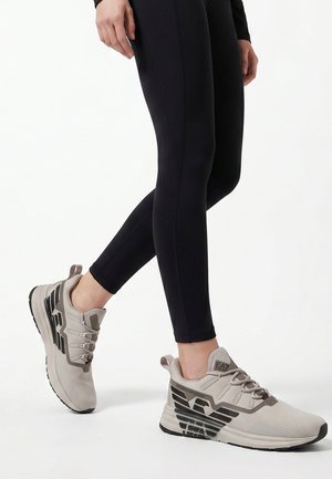 Persoon draagt grijze sportsneakers met een zwart logo en zwarte leggings, staand tegen een witte achtergrond.
