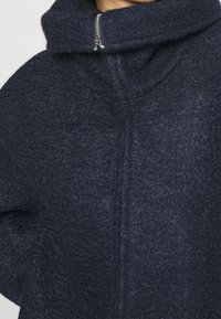 Mörkblå ullblandad jacka med hög krage, med blixtlås, strukturerad tyg och en avslappnad passform. Enkelt och modernt utseende.