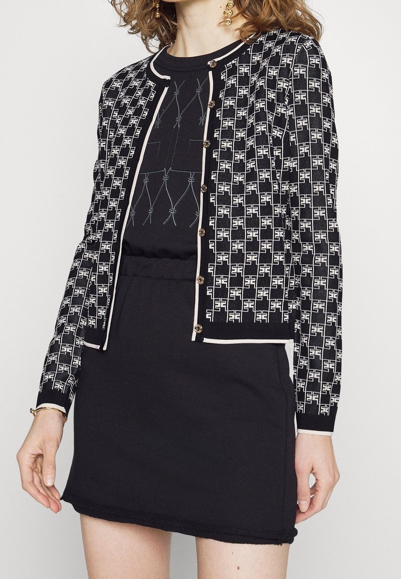 Cardigan a righe in bianco e nero con un tessuto testurizzato, scollatura rotonda, rifiniture a contrasto e bottoni dorati, abbinato a un semplice abito nero.